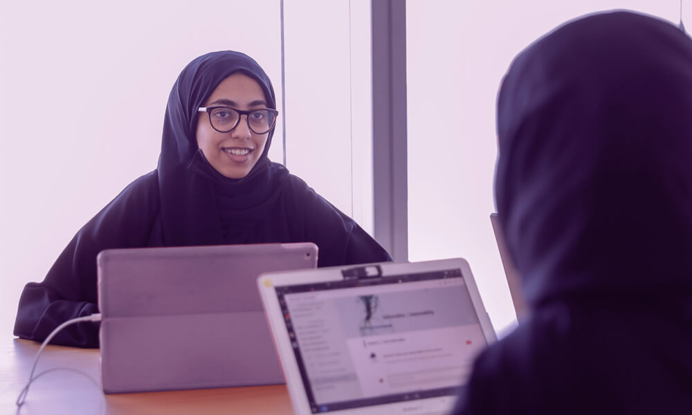 Two Arab women students sitting at a table with laptops engaged in a conversation - AGF a Philanthropic organization in UAE