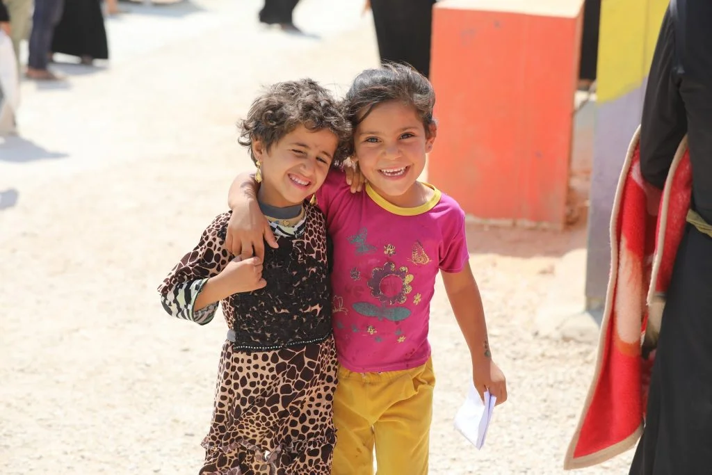 Two smiling children stand close together outdoors in Gaza. One child is holding a piece of paper.