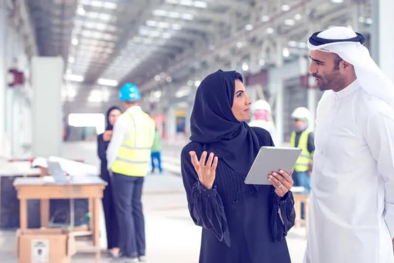 Two architects in traditional Emirati clothes standing on a construction site and talking. Woman is holding digital tablet. They look happy with results. There are some construction workers in back.