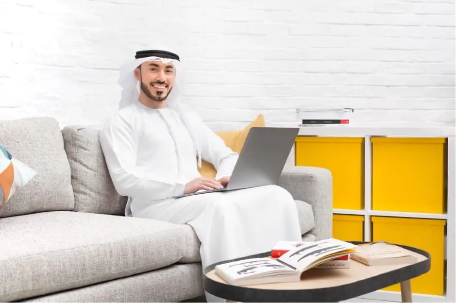Emirati man in traditional attire smiling at the camera while using a laptop on a gray sofa.
