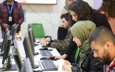 Students work at desktop computers in a computer lab, while one person stands and leans over another to offer guidance.