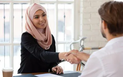 Two individuals are seated at a table, shaking hands with each other, possibly in a business meeting