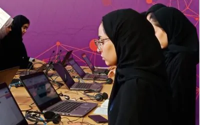 Women students from AGF attend an online class at a long table, using laptops with headsets placed nearby.