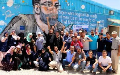 A diverse group of AGF members poses together in front of a large mural in Gaza, gathered to support relief efforts