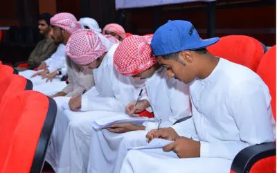 Young Arab students in traditional attire sit and focus on writing in their notebooks during a workshop.