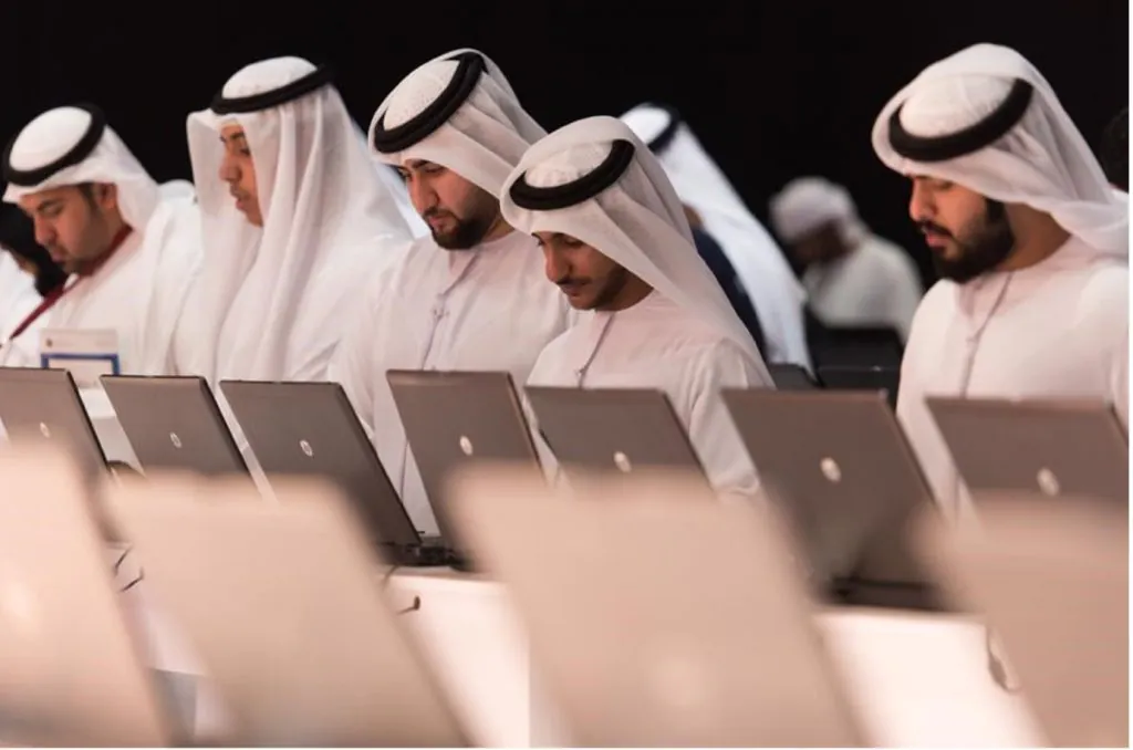 A group of men dressed in traditional Middle Eastern attire sit together, working on their laptops.