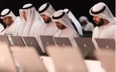 A group of men dressed in traditional Middle Eastern attire sit together, working on their laptops.