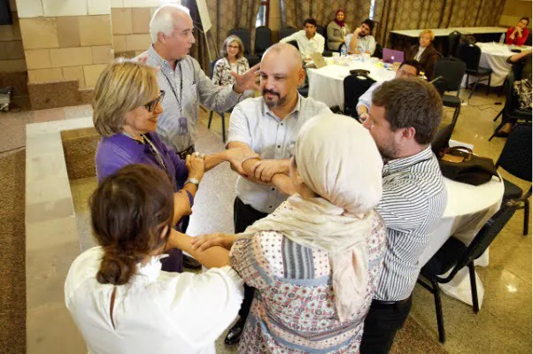 Six individuals stand in a circle, interlocking their arms and hands in a group activity