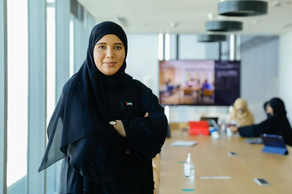 An Arab woman stands confidently in a modern conference room, wearing a small UAE flag pin with her arms crossed.