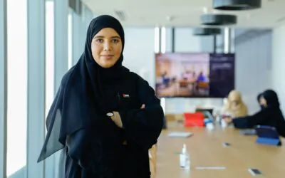 An Arab woman stands confidently in a modern conference room, wearing a small UAE flag pin with her arms crossed.