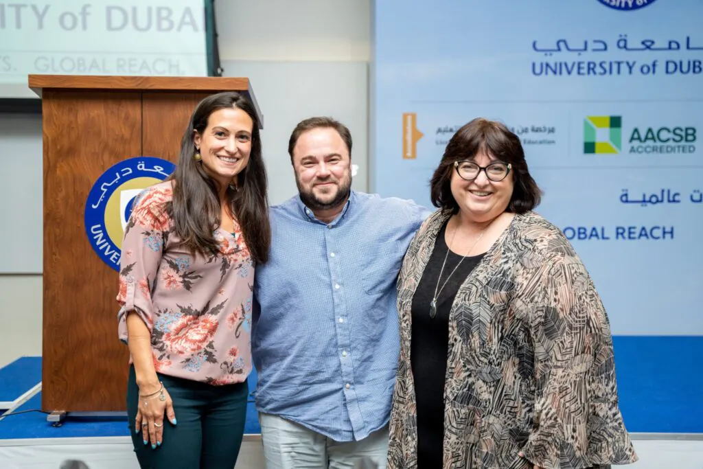 Three professional stand together, smiling at the camera in front of a banner displaying the University of Dubai logo.