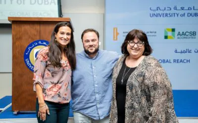 Three professional stand together, smiling at the camera in front of a banner displaying the University of Dubai logo.