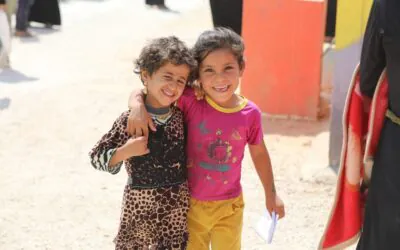 Two smiling children stand close together outdoors in Gaza. One child is holding a piece of paper.